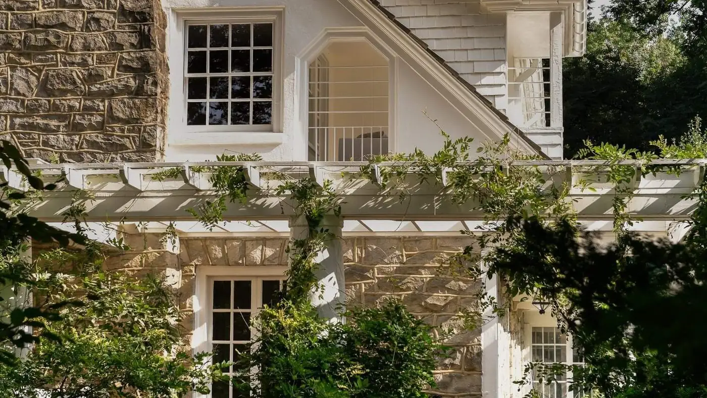 A stone and stucco house with a white pergola and wisteria in afternoon light
