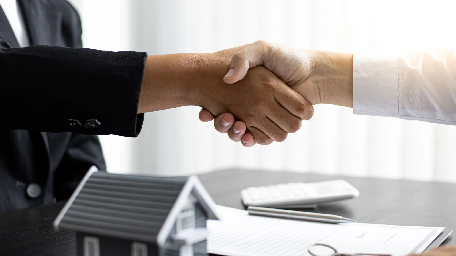 A handshake over a closing table with a model house, calculator, pen, and contract